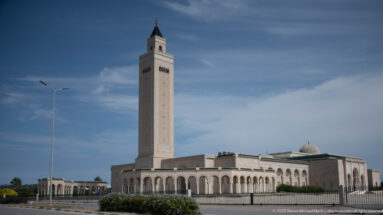 Roadside view of Malik ibn Anas Mosque with square minaret, dome, long arcades, fence, and streetlight.