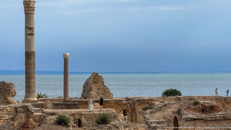 Two tall stone columns rising above the ruins of the Baths of Antoninus with the Mediterranean behind.
