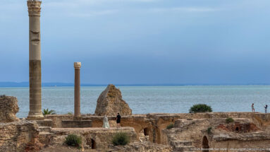 Two tall stone columns rising above the ruins of the Baths of Antoninus with the Mediterranean behind.