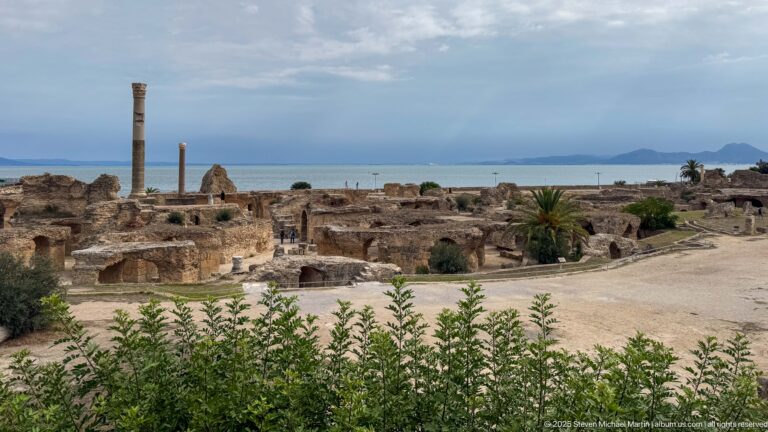 Broad panorama of the Baths of Antoninus showing vaulted ruins, columns, Mediterranean water, and distant hills.