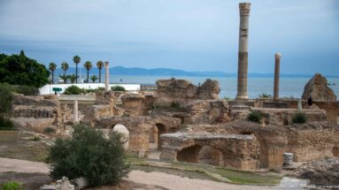 Vaulted Roman bath foundations and tall columns at the Baths of Antoninus, with palms and Mediterranean beyond.
