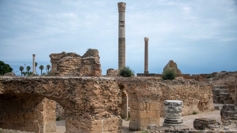 Two tall columns above vaulted ruins of the Baths of Antoninus with a carved capital fragment in the foreground.