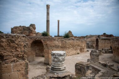 Large weathered Corinthian-style capital fragment among vaulted ruins and standing columns of the Baths of Antoninus.