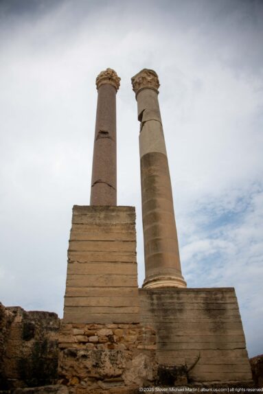 Two tall stone columns rising from a stepped base at the Baths of Antoninus against a cloudy sky.