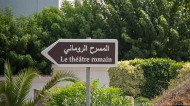Brown directional sign in Arabic and French pointing left toward the Roman Theatre amid green landscaping.