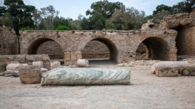 Three masonry arches behind a fallen veined marble column shaft and scattered ancient stone blocks.