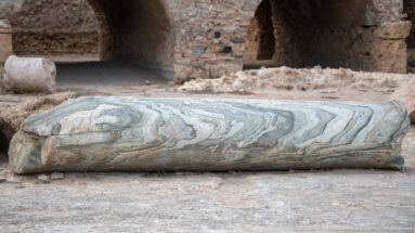 Close view of a fallen pale marble column shaft with pronounced gray-green veining and chipped edges.