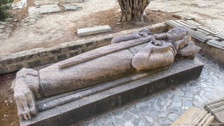 Oblique overhead view of a robed recumbent stone effigy on a dark rectangular tomb base.