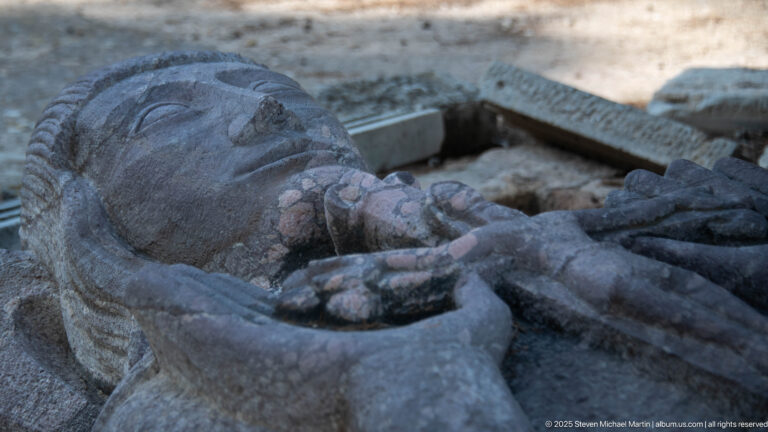 Close view of the weathered face and folded hands of a recumbent stone clerical effigy.