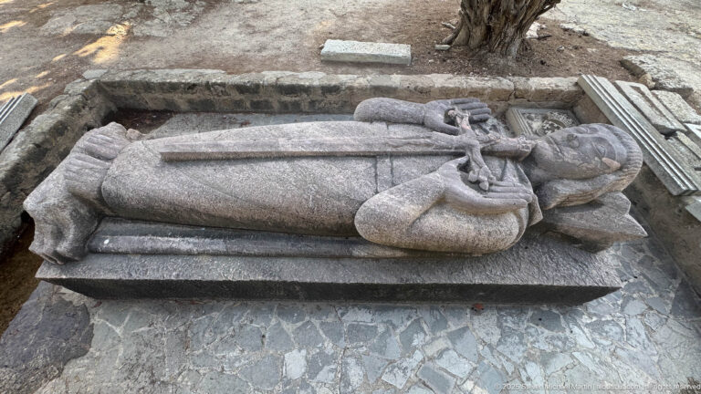 Three-quarter overhead view of a full recumbent stone clerical effigy on a dark tomb base.