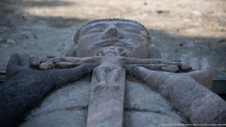 Low lengthwise view along a recumbent stone effigy, with robe folds leading toward the face and hands.