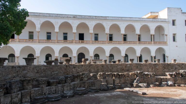 Excavated stone foundations in front of a two-story white building with repeated rounded arcades on Byrsa Hill.
