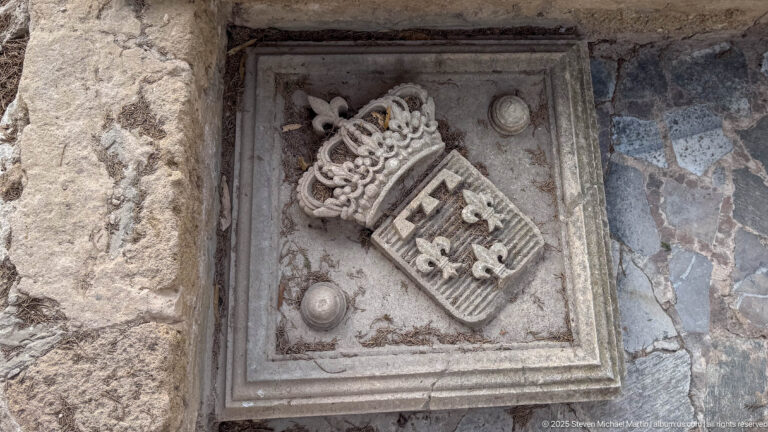 Weathered square stone plaque carved with an ornate crown above a heraldic shield and small surrounding emblems.