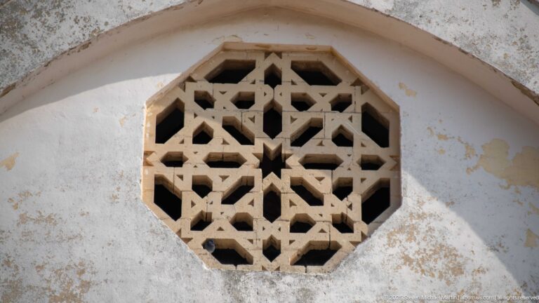 Octagonal pierced stone window with interlaced geometric tracery and a small central star in a weathered pale wall.
