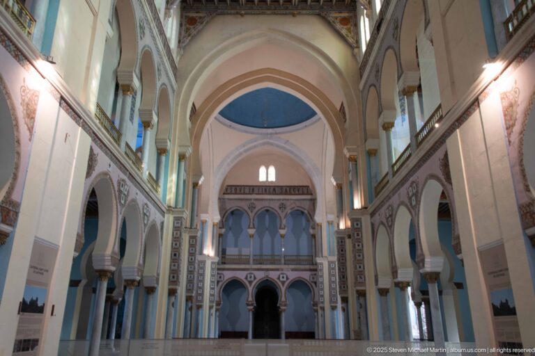 Long interior view through the nave of Carthage L’Acropolium with tall arches, galleries, painted columns, and blue vaulted bays.