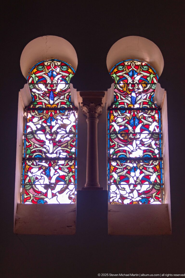 Pair of tall multicolored stained-glass windows with red, blue, yellow, and green patterns separated by a slender column.