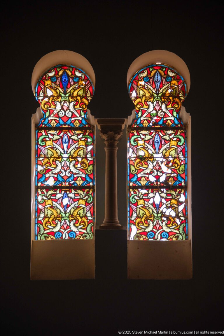 Symmetrical pair of richly colored stained-glass windows with red, yellow, blue, and green ornamental patterns and a central column.