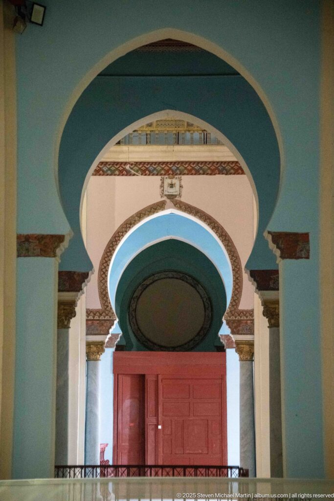 Nested blue, white, and pink arches frame a red wooden doorway inside Carthage L’Acropolium.
