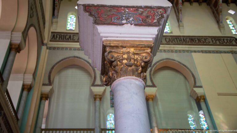 Gilded Corinthian-style capital on a pale column beneath painted arches and Latin inscription bands.