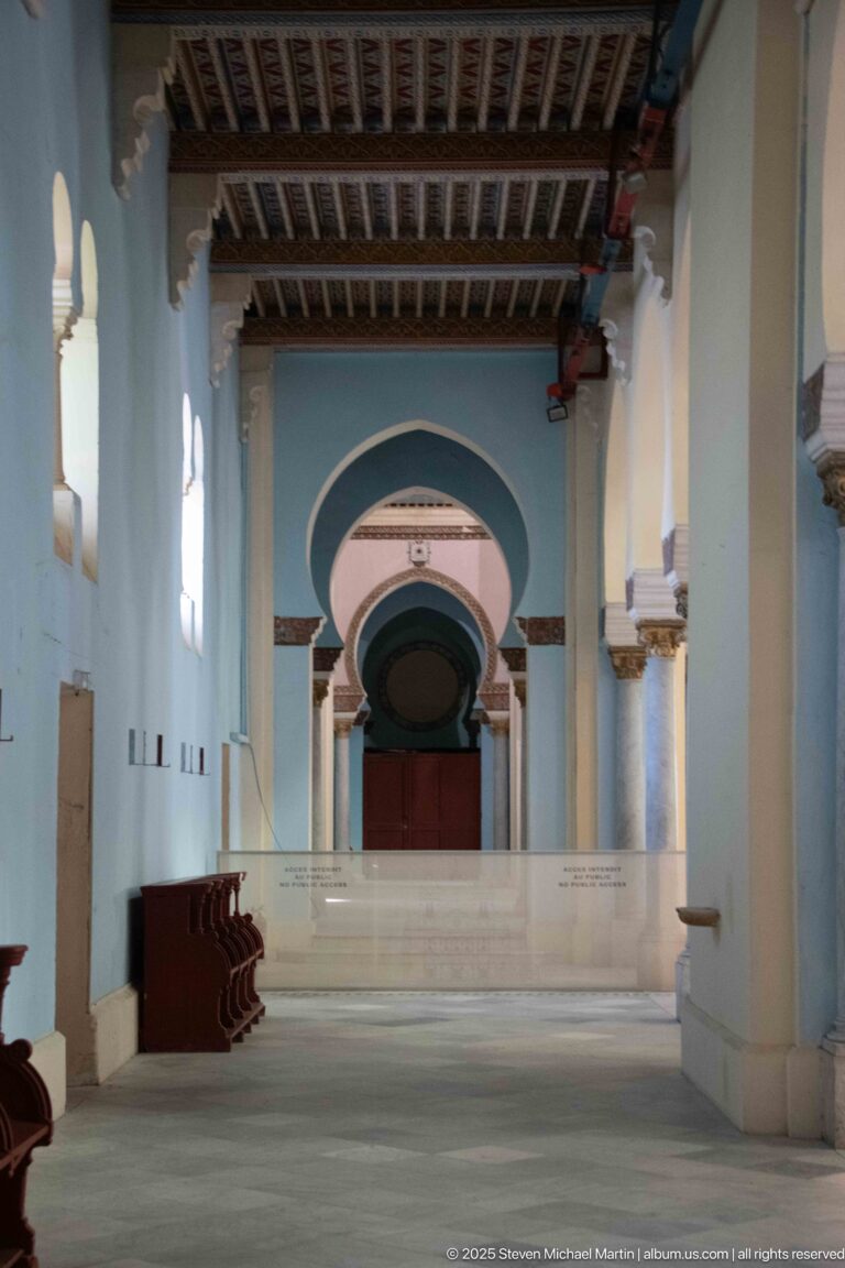 Long side aisle with repeated columns, blue-green arches, wooden ceiling, and a red door at the far end.