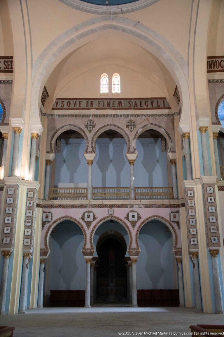 Two-level painted arcade beneath a Latin inscription band, with pale columns and muted blue wall panels.