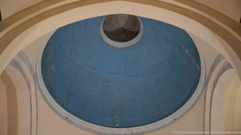 Upward view into a blue-painted dome with a circular opening at the crown, pale ribs, and visible cracks.