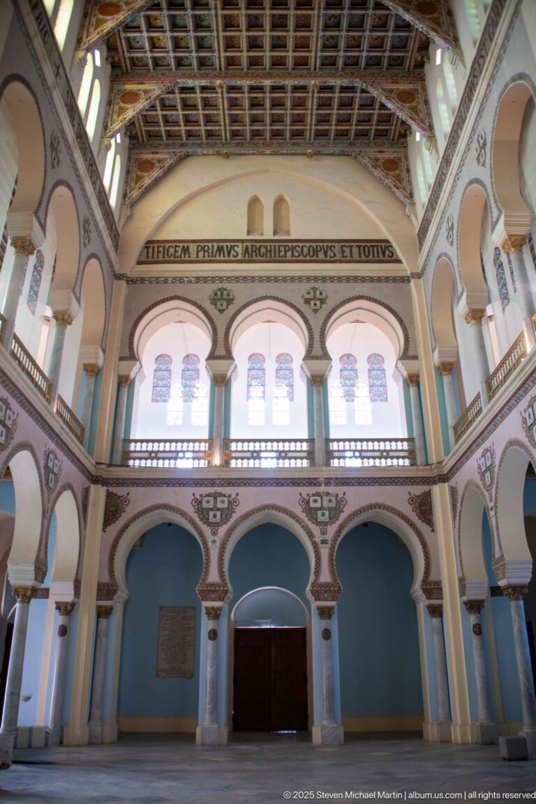 Wide upper interior with three stained-glass window bays, gallery railing, rounded arches, Latin inscription, and patterned ceiling.