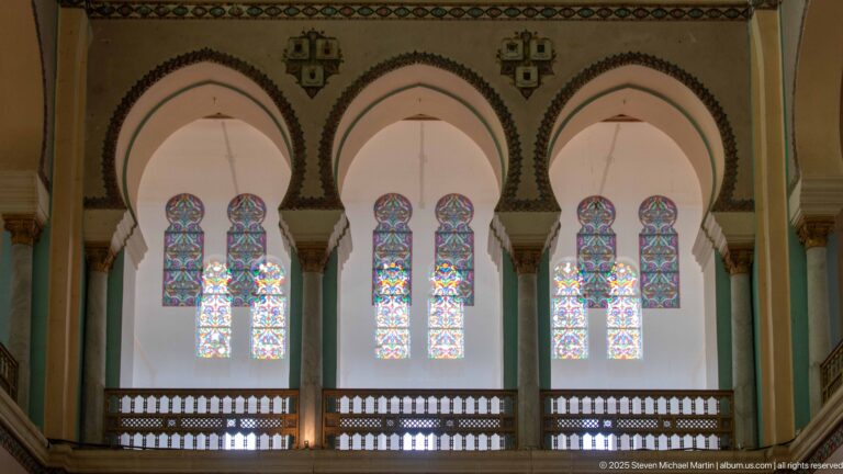 Six tall stained-glass windows grouped in pairs beneath three rounded arches above an interior gallery railing.