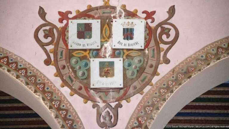 Painted heraldic wall composition with three labeled shields surrounded by curling red, green, and gold ornament.