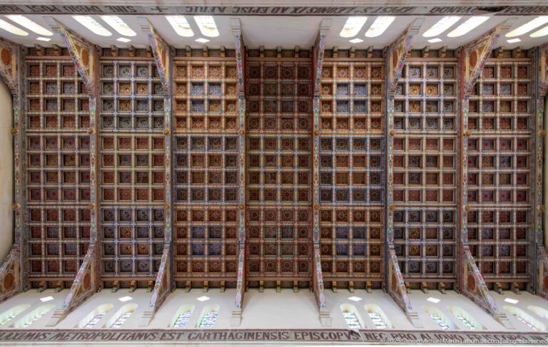 Upward view of a large compartmented wooden ceiling covered with dense red, gold, blue, and dark geometric patterns.
