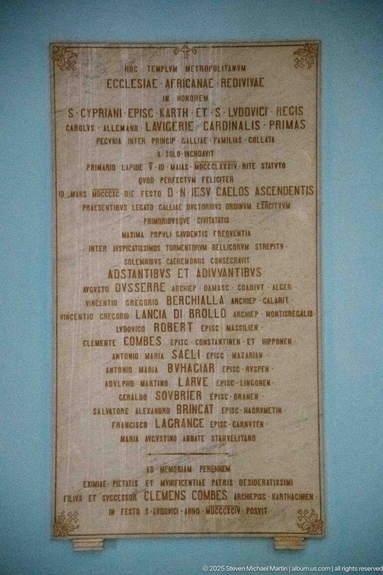 Large rectangular Latin inscription plaque with centered lines of text and prominent ecclesiastical names on a blue wall.