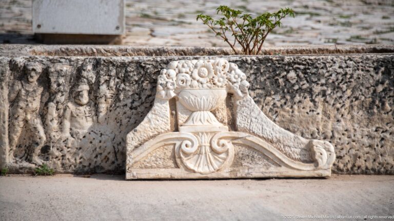 Carved stone architectural fragment with paired volutes, central fan-shaped ornament, and bead-like decoration against rough masonry.