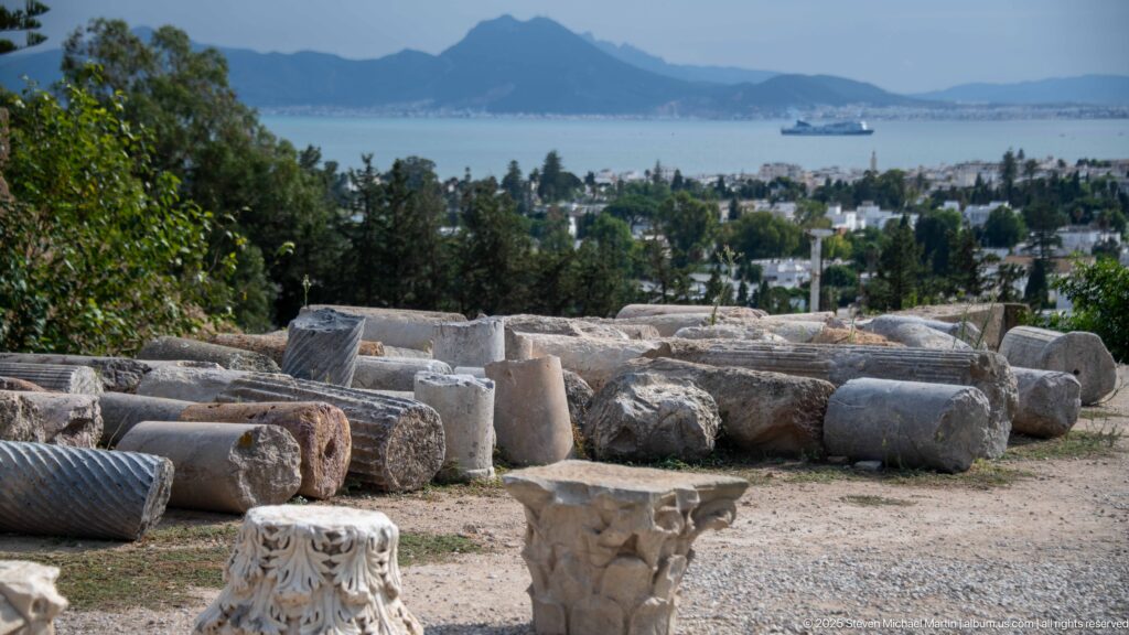 Scattered column shafts and carved stone fragments on Byrsa Hill overlooking the Gulf of Tunis and distant mountains.