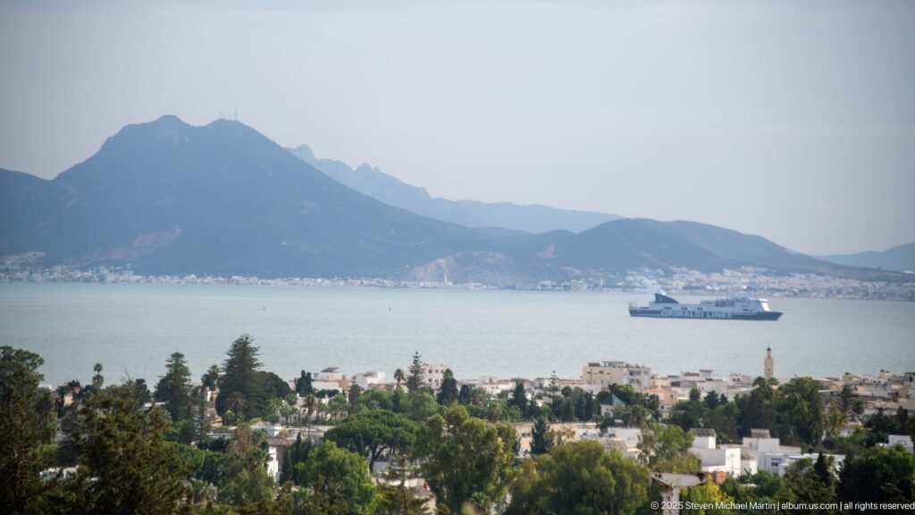 View from Byrsa Hill over trees and shoreline to the Gulf of Tunis, a ship, and hazy mountains beyond.