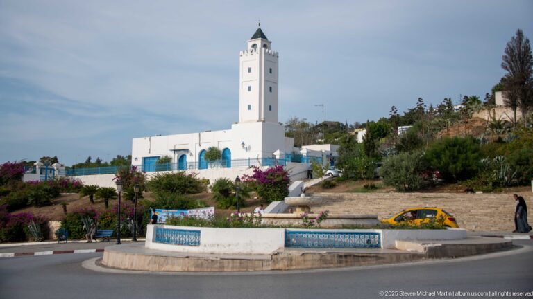 White mosque with tall square minaret behind a landscaped roundabout, flowering shrubs, palms, pedestrians, and a yellow taxi.