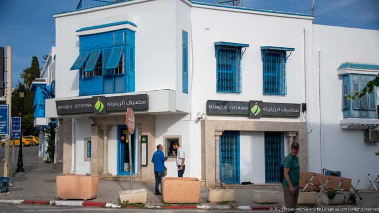 Banque Zitouna branch with Arabic and Latin signage on a white building with blue doors, grilles, and projecting upper window.