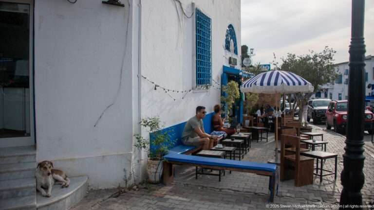 Blue café tables and chairs beside a white wall, striped umbrella and patrons farther down the lane, with a cat on nearby steps.