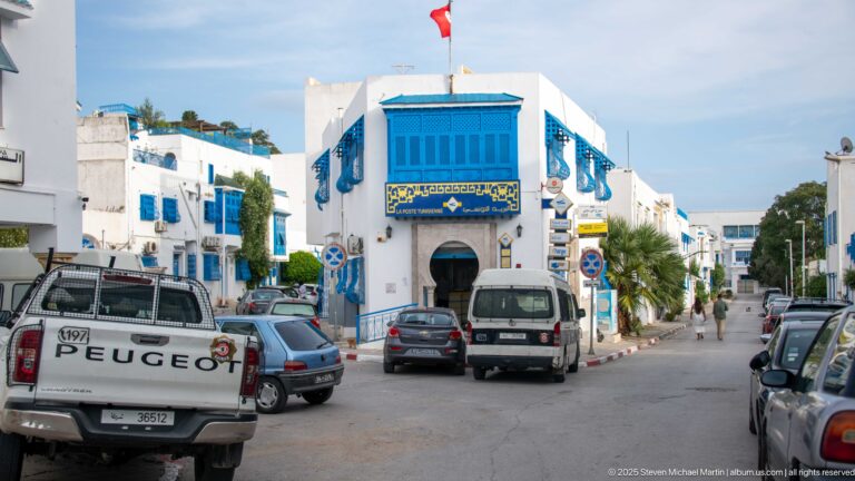 White corner building with bright blue projecting window and shop signs, surrounded by cars and vans on a Sidi Bou Said street.