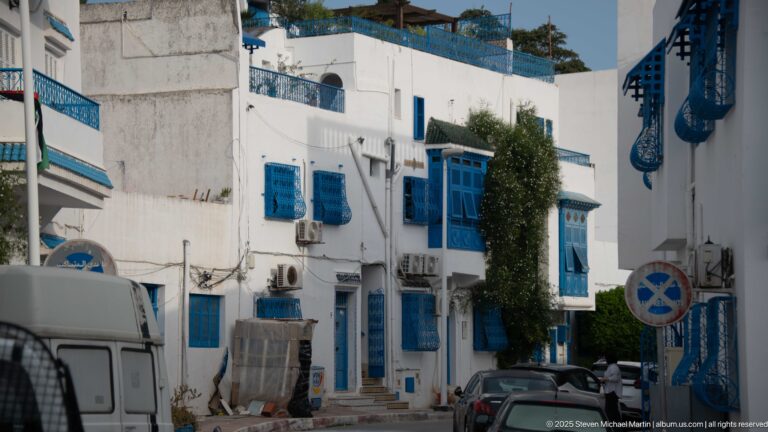 Street lined with multi-story white buildings, blue projecting windows and shutters, parked vehicles, and decorative blue brackets.