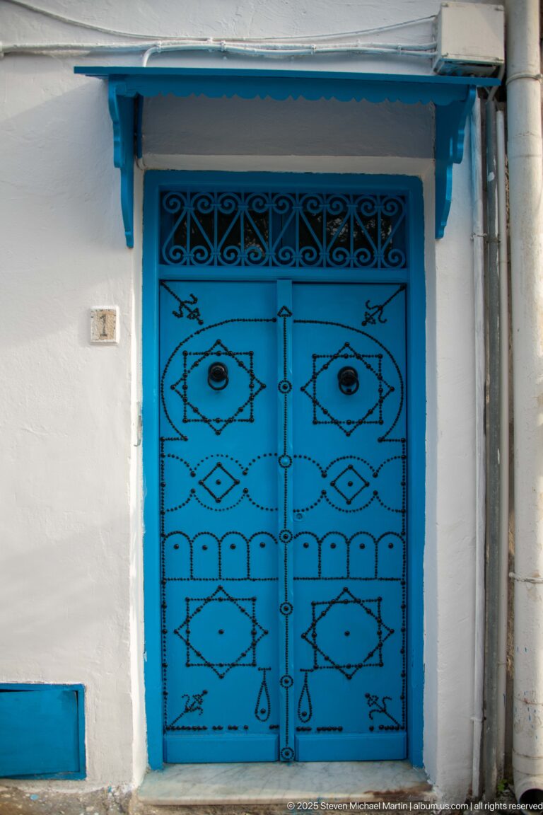Blue double metal door with symmetrical black geometric designs, topped by a narrow grille and blue awning in a white wall.