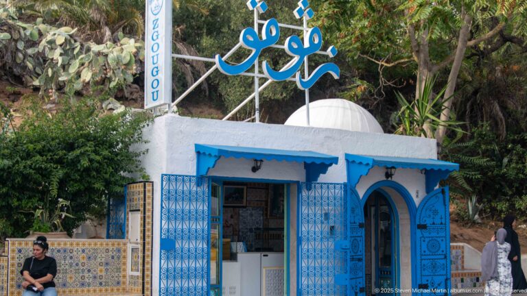 Small white shop with blue trim and open gates, patterned tile panels, Arabic roof lettering, and dense greenery behind it.