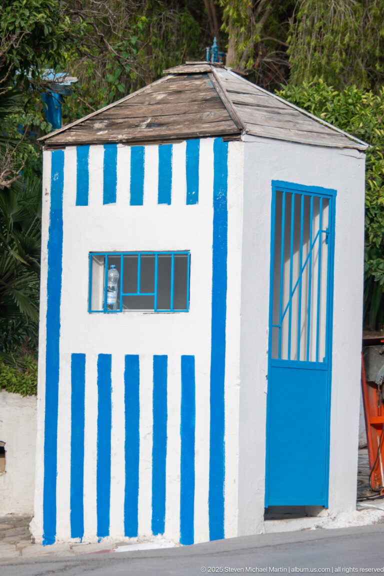 Small white roadside hut with vertical blue stripes, a blue-framed window, barred blue door, and low sloping roof.
