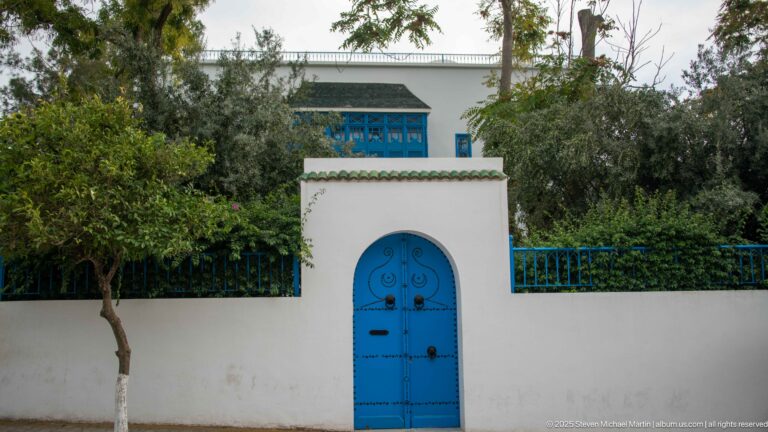 Arched blue metal gate centered in a high white wall, with blue railings, trees, and part of a residence visible above.