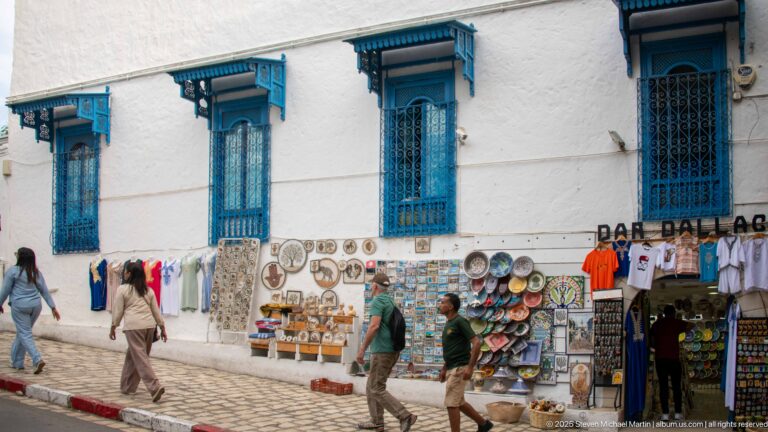 Souvenir displays of ceramics, plates, clothing, and keepsakes beneath tall blue-grilled windows as pedestrians walk along the cobbled street.