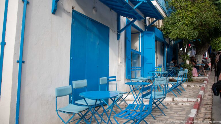 Empty blue folding chairs and round café tables beside a white building with blue doors along a narrow cobbled lane.