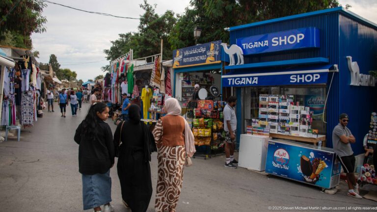 Blue Tigana Shop kiosk beside a crowded pedestrian lane with souvenir stalls, hanging clothing, merchandise, and visitors.