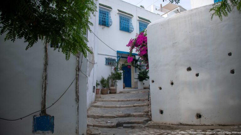 Narrow stepped lane between white walls leading toward a blue door and blue-grilled windows beneath magenta bougainvillea.