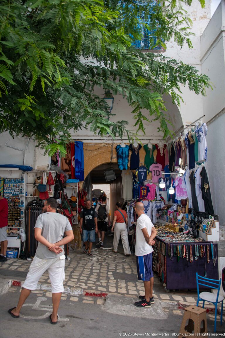 Shaded market entrance with clothing, sports jerseys, jewelry and souvenir displays as visitors walk beneath leafy trees.