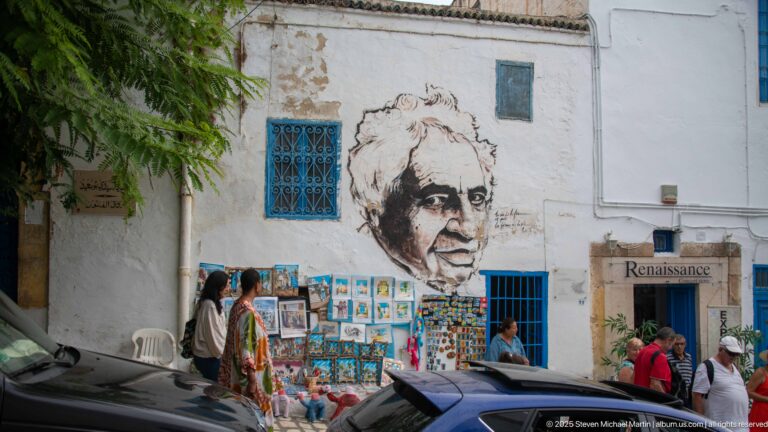 Large black-and-white portrait mural on a white wall above colorful street art, with blue window grilles and a “Renaissance” sign nearby.