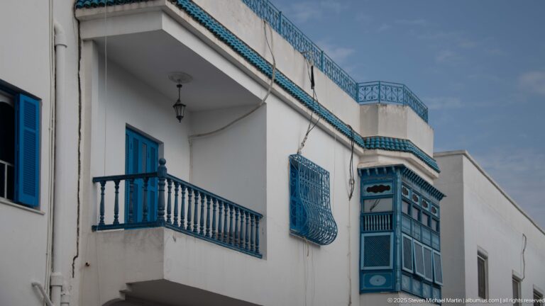 Upper story of a white house with blue balcony railings, projecting window grilles, enclosed bay, roof trim, and a hanging lantern.